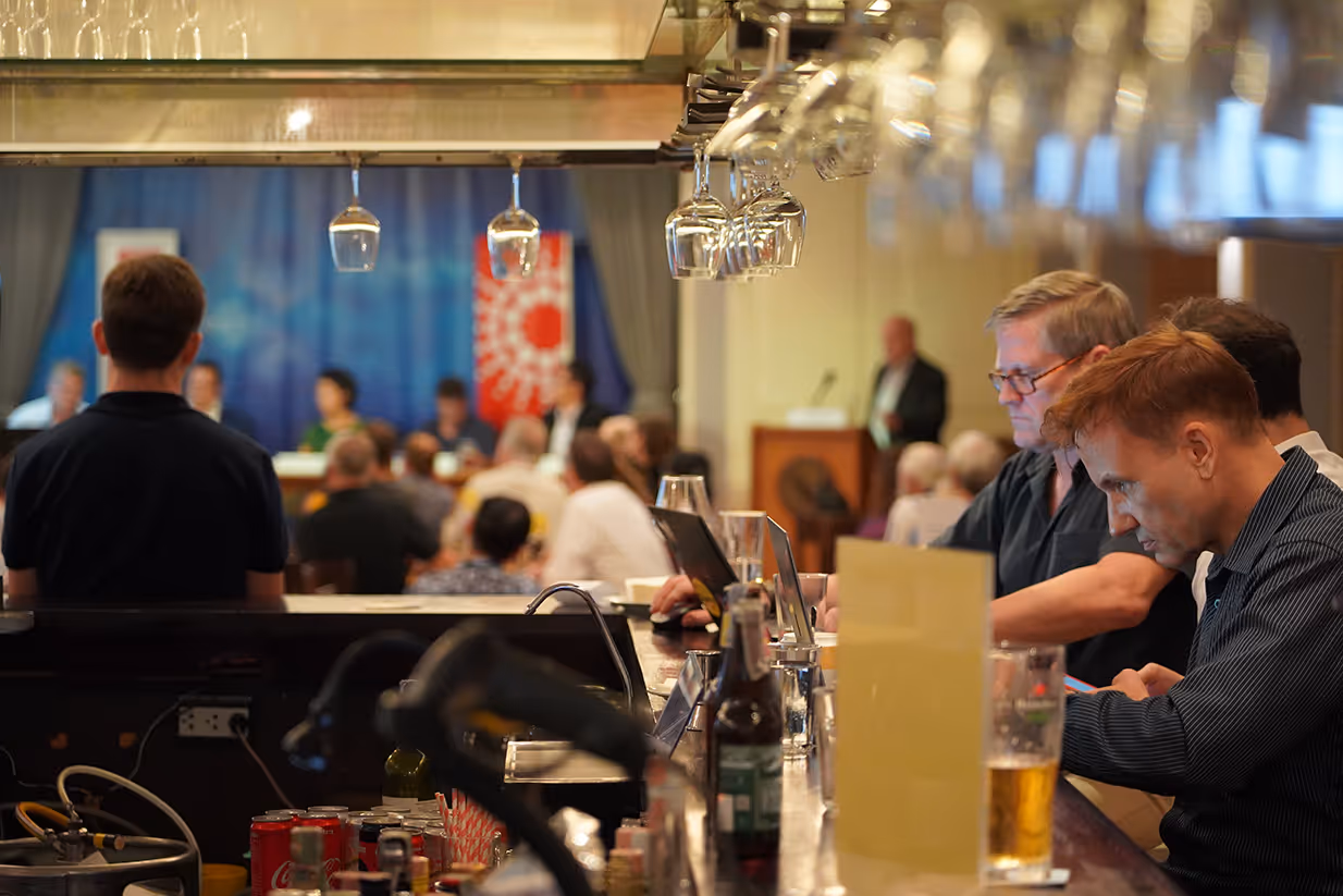 Members at the bar with drinks in the foreground, an audience seated facing the lit stage behind
