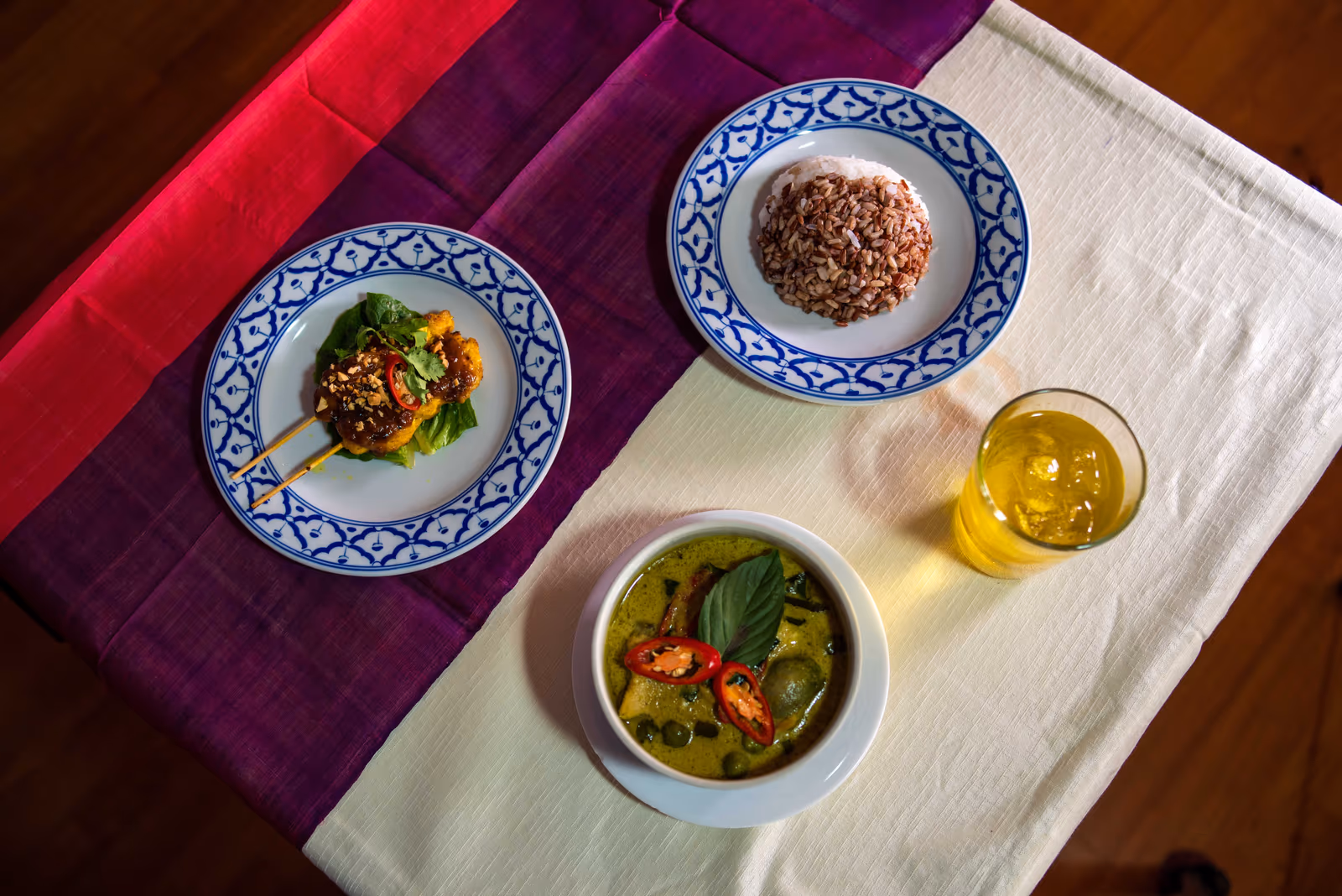Two plates of Thai food and a bowl of green curry with a glass of tea, seen from above