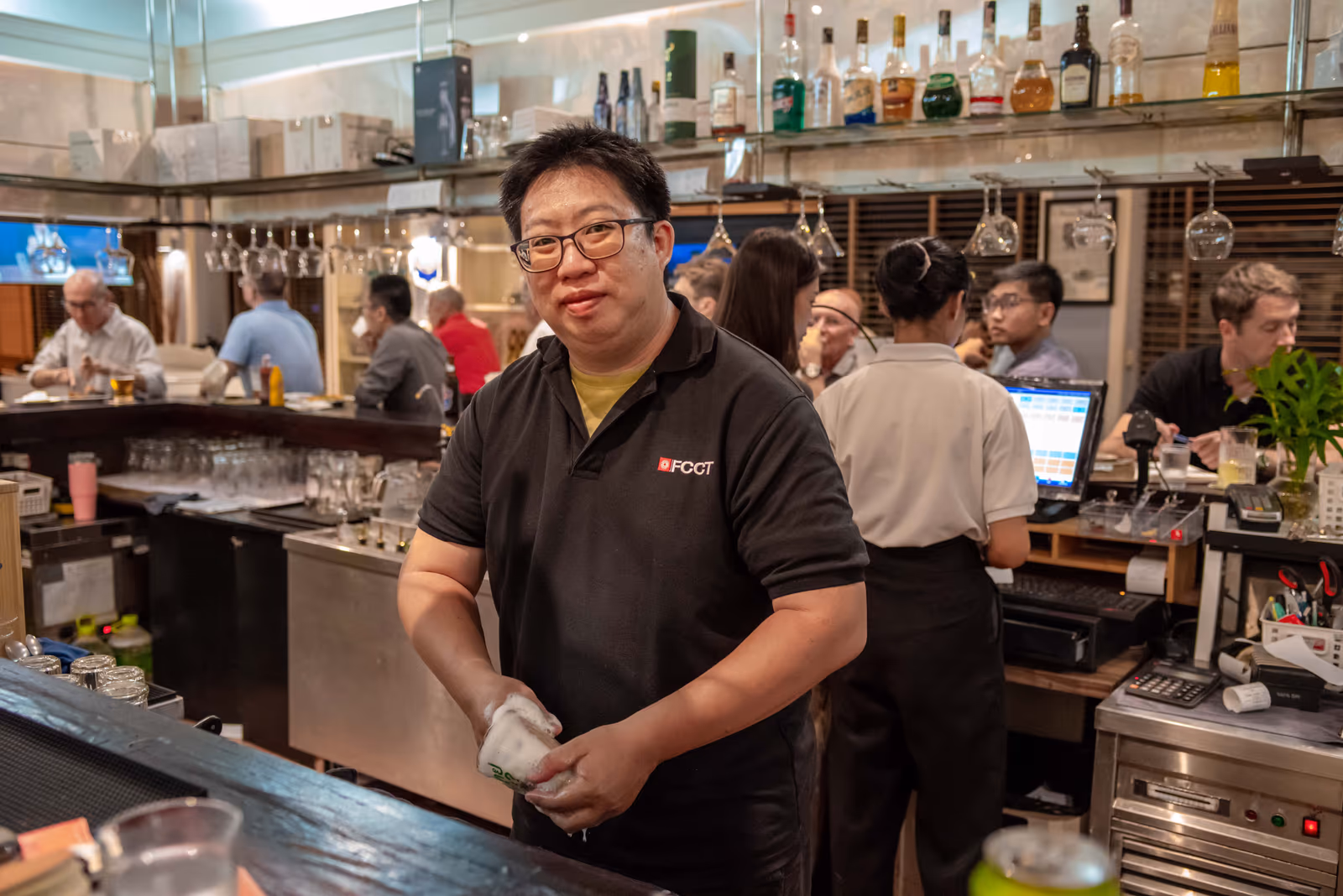 A member of bar staff in an FCCT shirt behind the bar, with guests and colleagues around them
